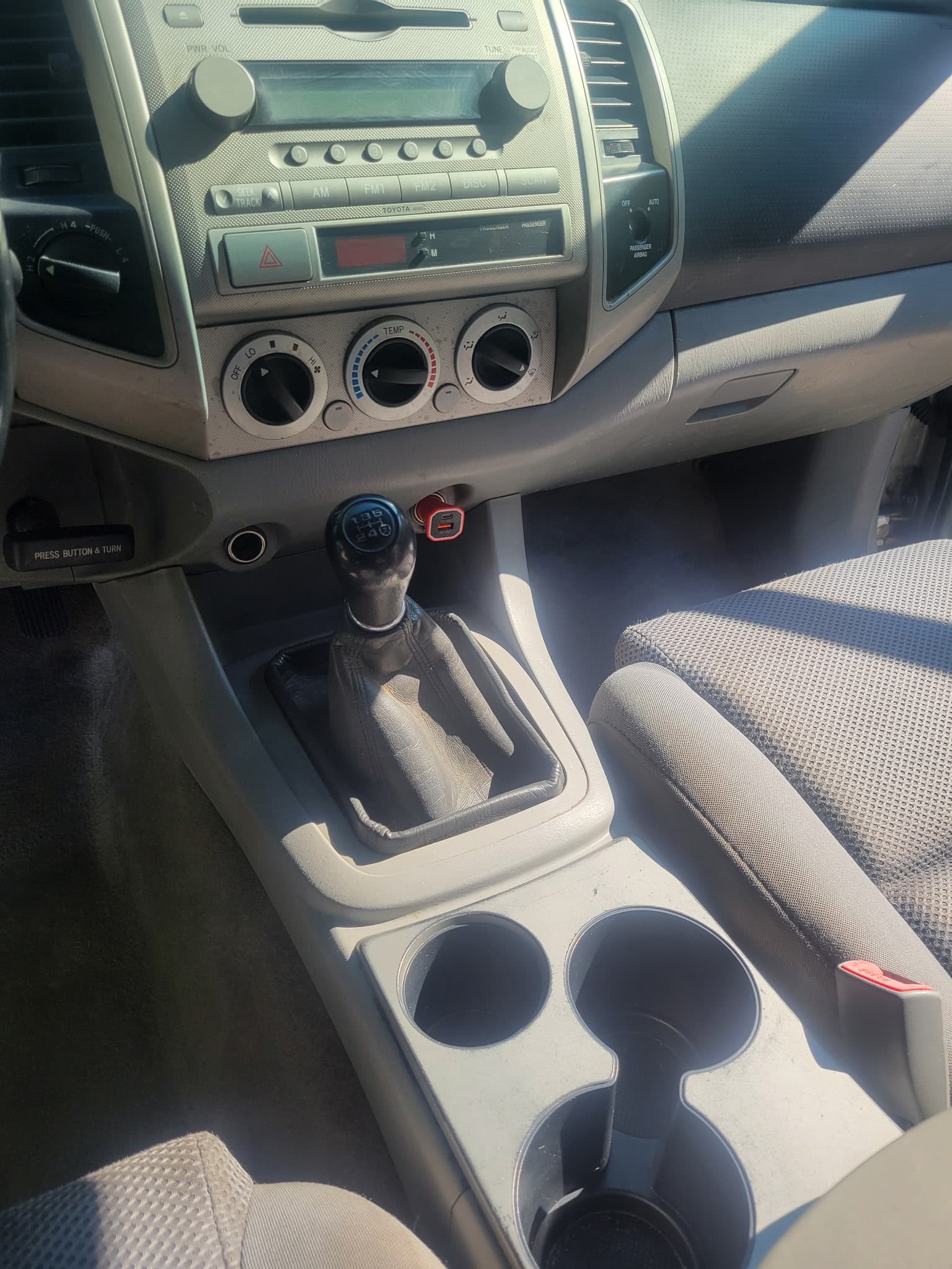 Interior of a vehicle featuring a manual gear shifter, climate controls, and cup holders.