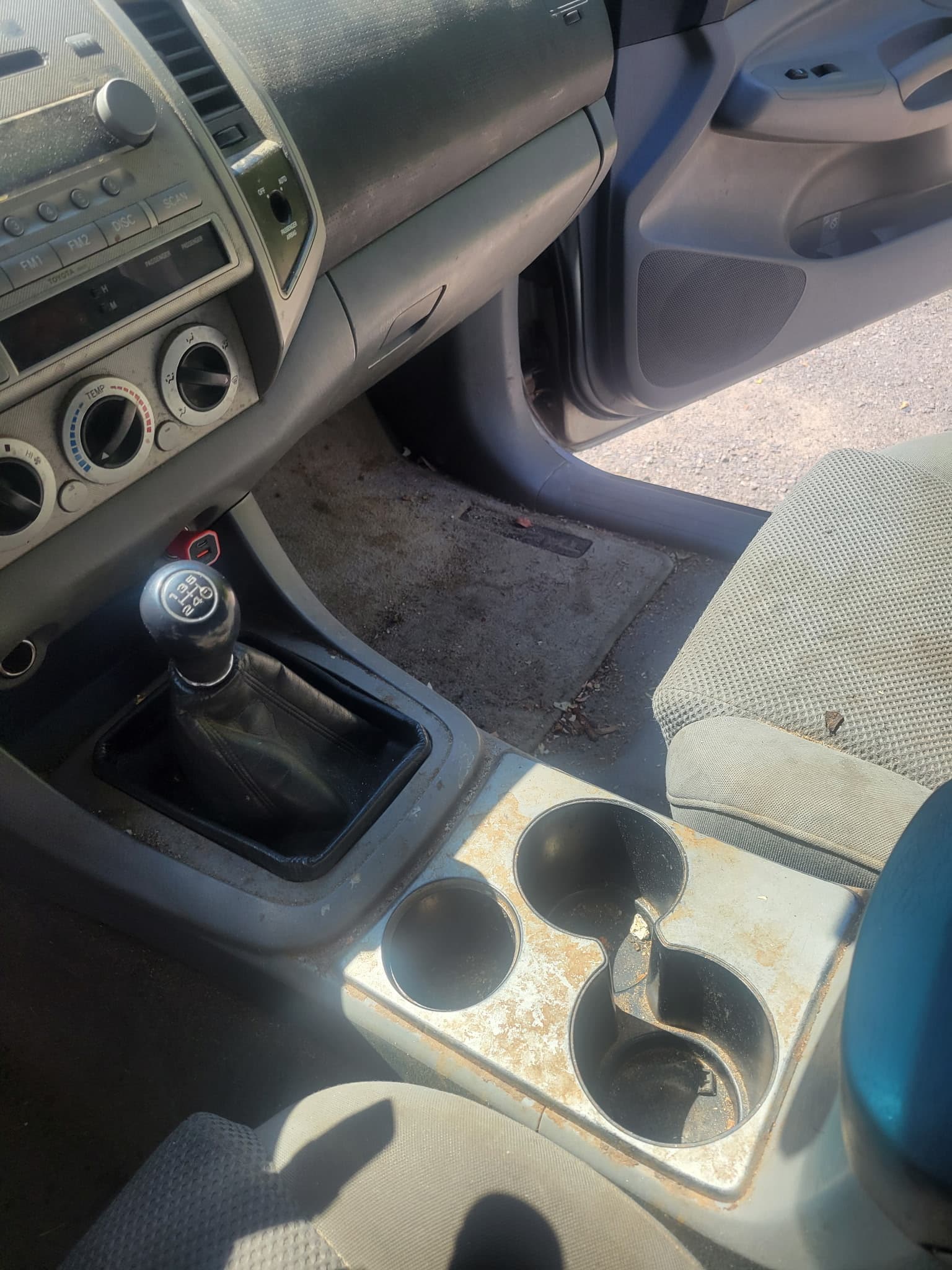 Dirty car interior with manual gear shifter, dusty dashboard, and stained center console cup holders.